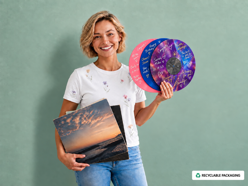 A smiling woman stands against a muted green wall holding vinyl records and a record sleeve. She wears a white graphic T-shirt and blue jeans.